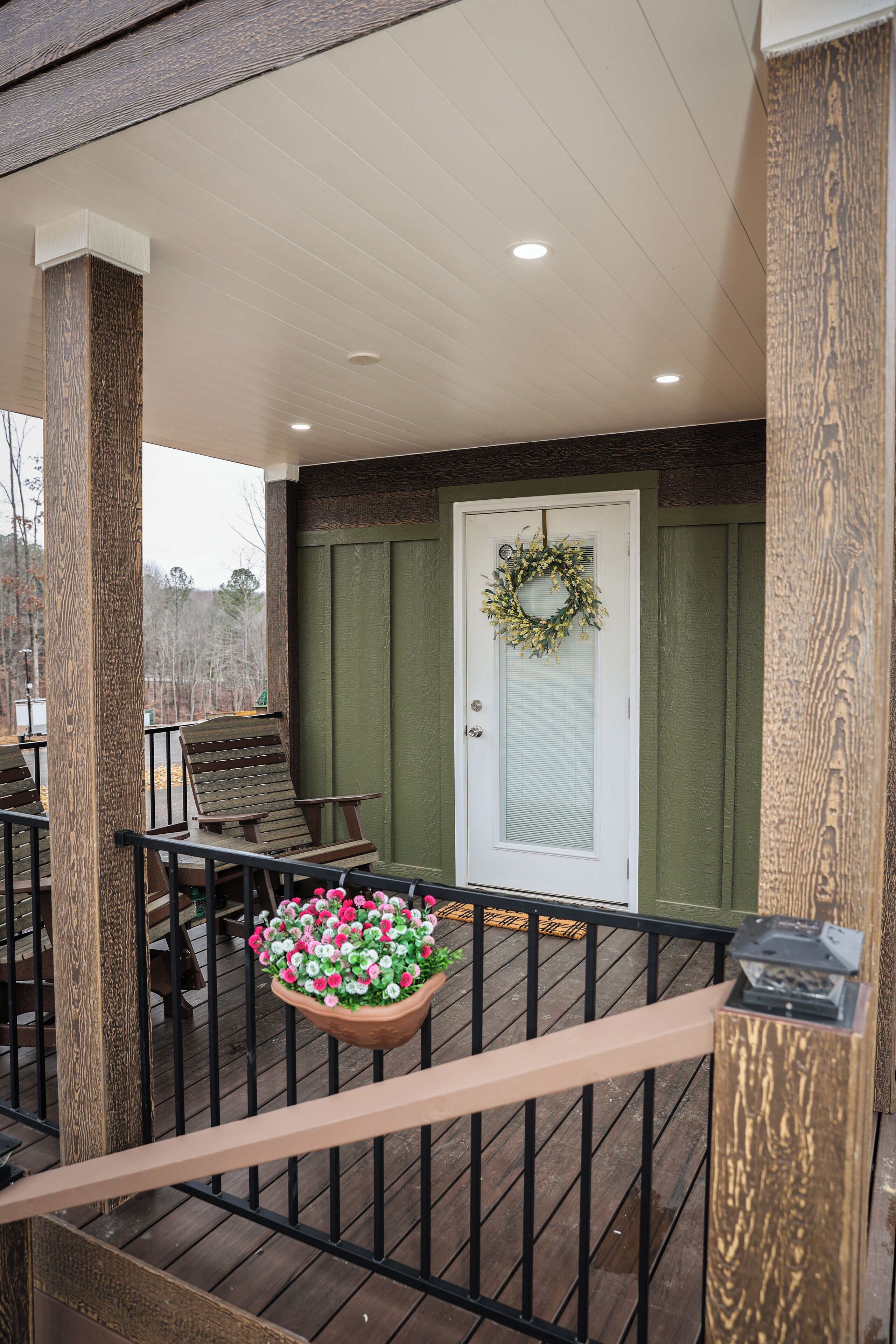 A covered porch with a wooden railing, a door with a wreath, and a planter with flowers.