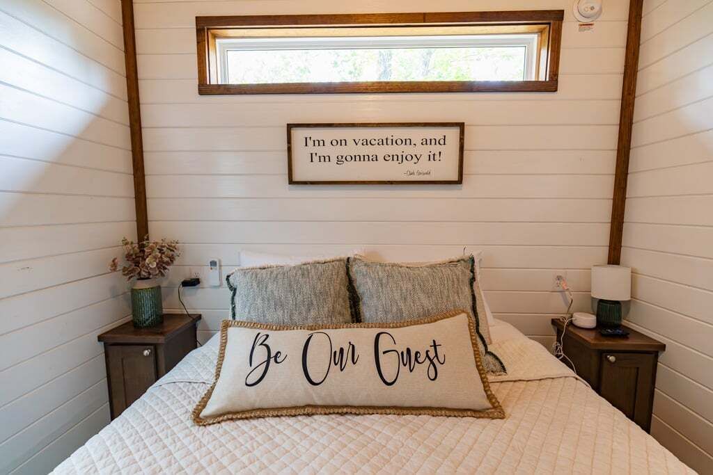 Un dormitorio con cama, dos almohadas, una mesita de noche, lámpara, una cita enmarcada en la pared y ventana.