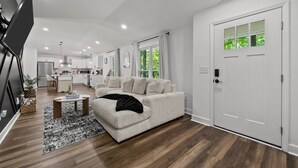 Ein modernes Wohnzimmer mit einer Couch, einem Couchtisch und einem Teppich. Im Hintergrund ist eine Küche mit Schränken und einem Kühlschrank zu sehen.