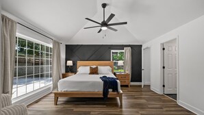 Ein Schlafzimmer mit einem großen Bett, einem Deckenventilator, einem Nachttisch und einem Fenster mit Vorhängen.