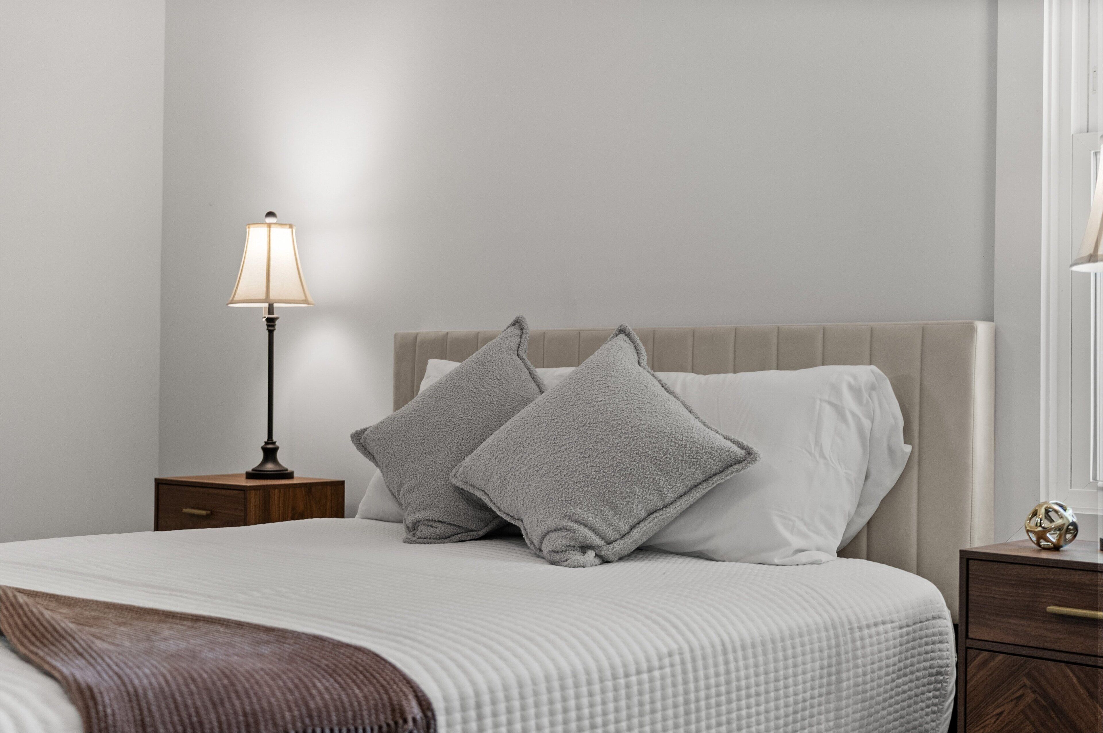 Un letto rifatto con un copriletto bianco e due cuscini grigi, una lampada da comodino e un comodino in legno con un cassetto.