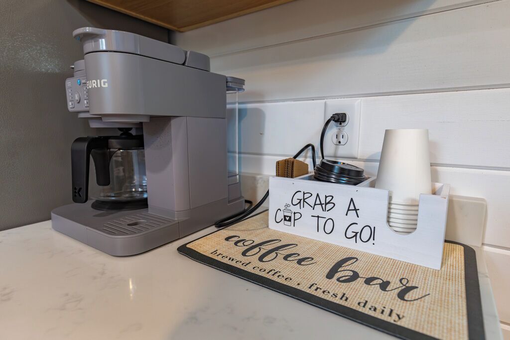 A coffee machine labeled "URIG" with a sign that reads "GRAB A CUP TO GO!" and a mat that says "coffee bar brewed coffee fresh daily".