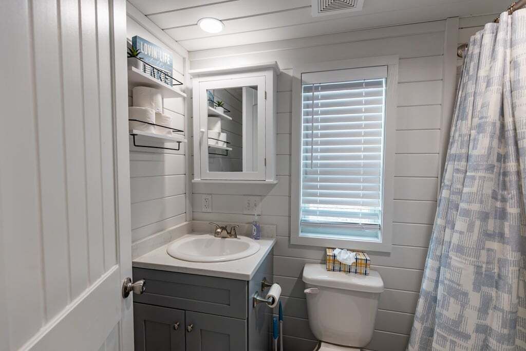 A bathroom with a white sink, a mirror above it, a toilet, and a window with blinds.