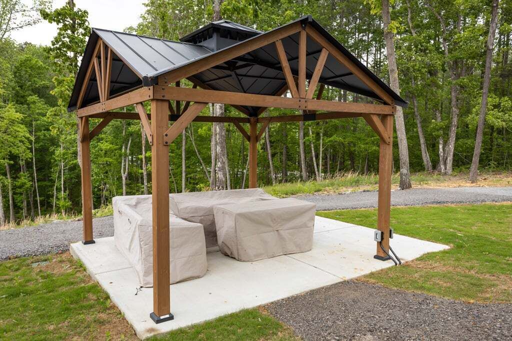 En trälusthus med ett tak av metall och beige kuddar på en betongplattform, omgivet av träd.