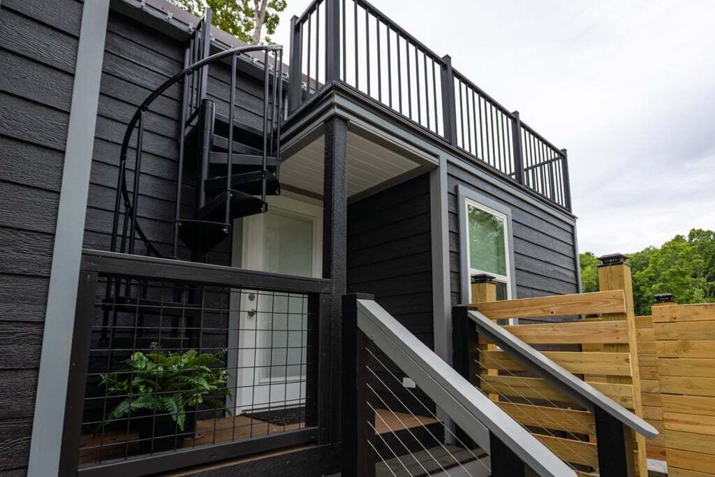 Un bâtiment à deux étages doté d’une terrasse en bois, d’un escalier en colimaçon et d’un espace clos agrémenté de plantes.
