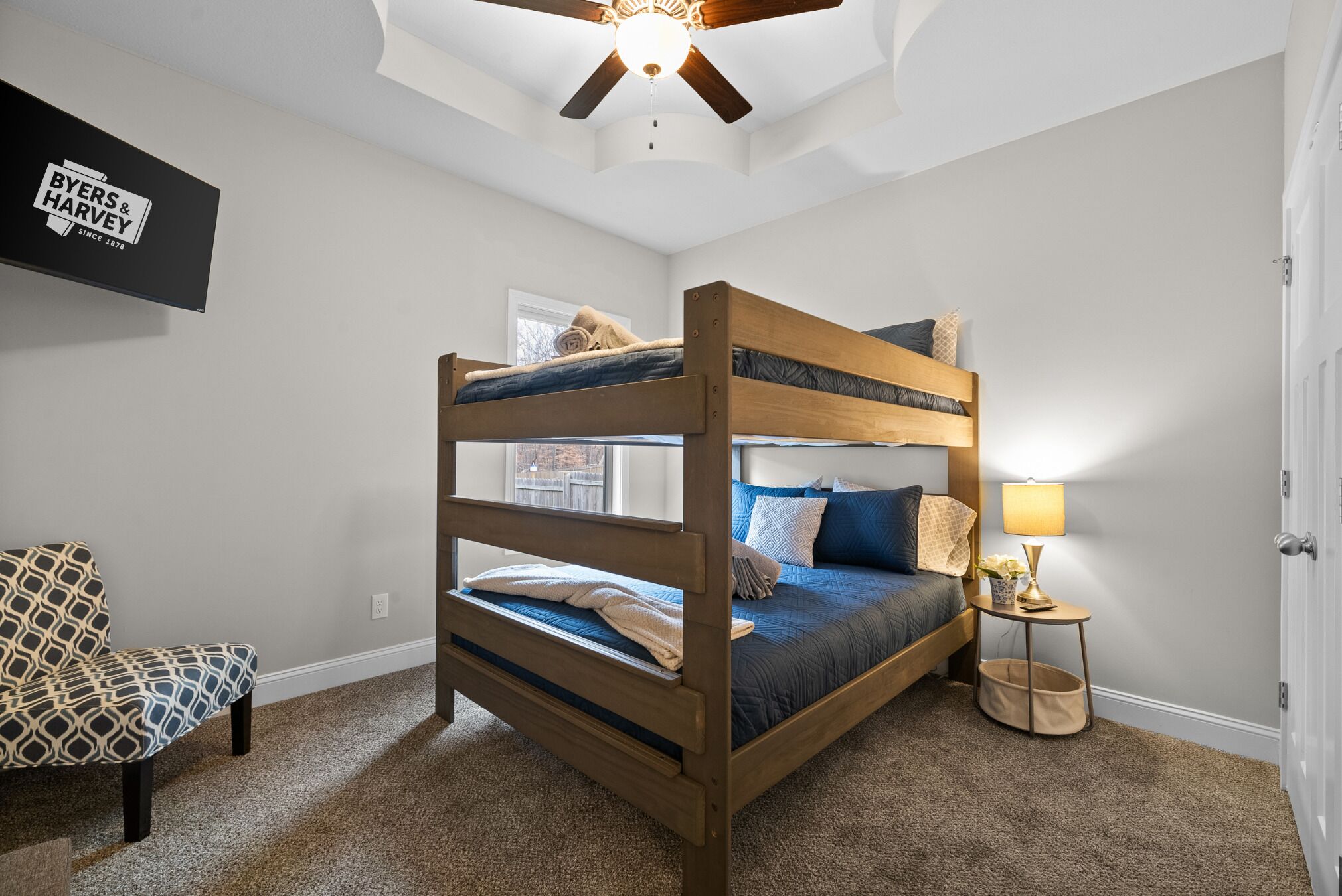 A bunk bed room with a ceiling fan, a TV mounted on the wall, a bedside table with a lamp, and a patterned chair.