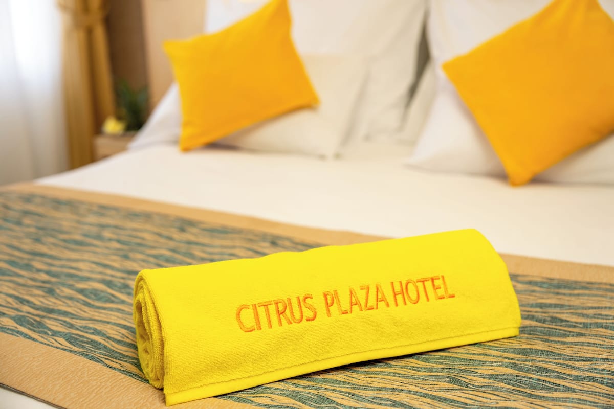 A neatly folded yellow towel with "CITRUS PLAZA HOTEL" printed on it, placed on a bed with white sheets and yellow pillows.