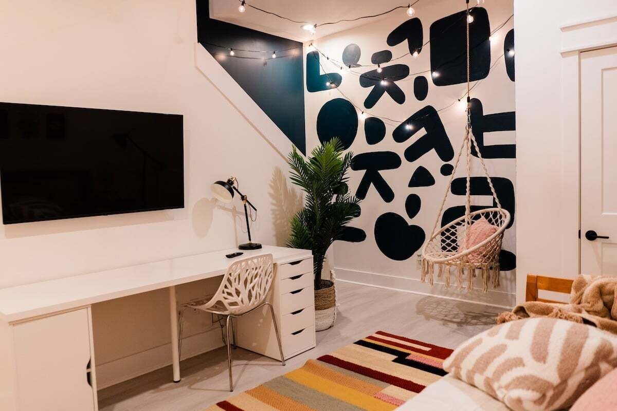 A modern bedroom with a white desk, a chair, a bed with a striped blanket, a hanging chair, and a wall with abstract black and white patterns.