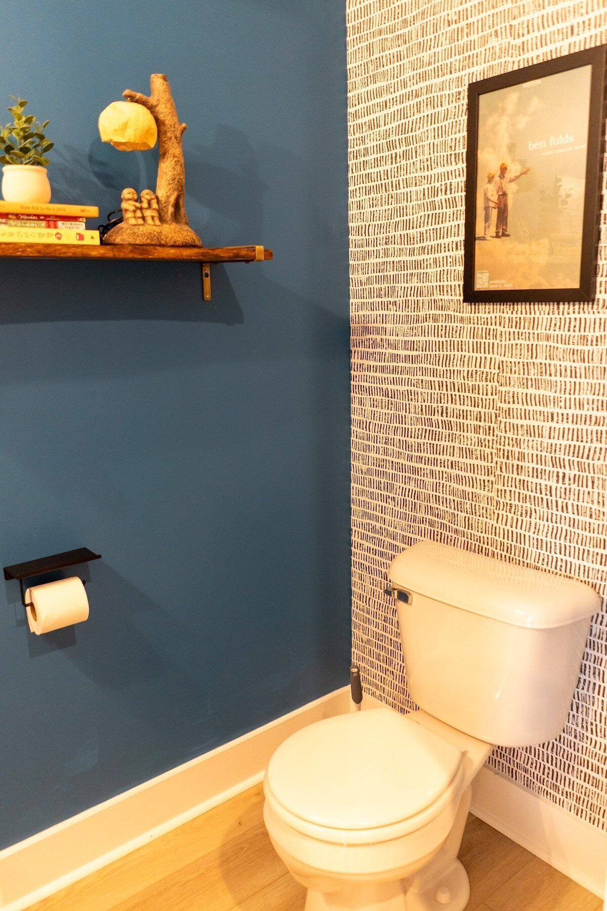 Un baño con pared azul, un inodoro, una estantería con libros y una planta, y un cuadro en la pared.