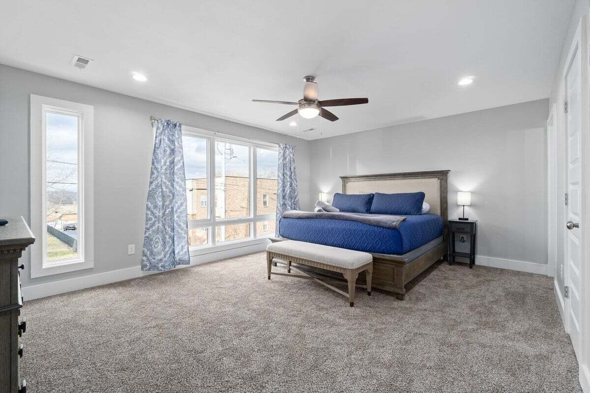 A bedroom with a bed, a bench, a nightstand, a ceiling fan, and large windows.