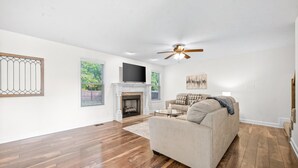 Ein geräumiges Wohnzimmer mit Holzdielen, einem beigen Sofa, einem Kamin und einem Deckenventilator.