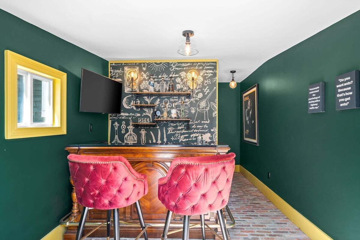 Un coin bar avec un mur recouvert d’un tableau noir, deux tabourets de bar rouges, un téléviseur et un sol carrelé imitant un mur de briques.