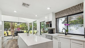 A modern kitchen with a large island, white cabinetry, and large windows offering views of the outdoors.