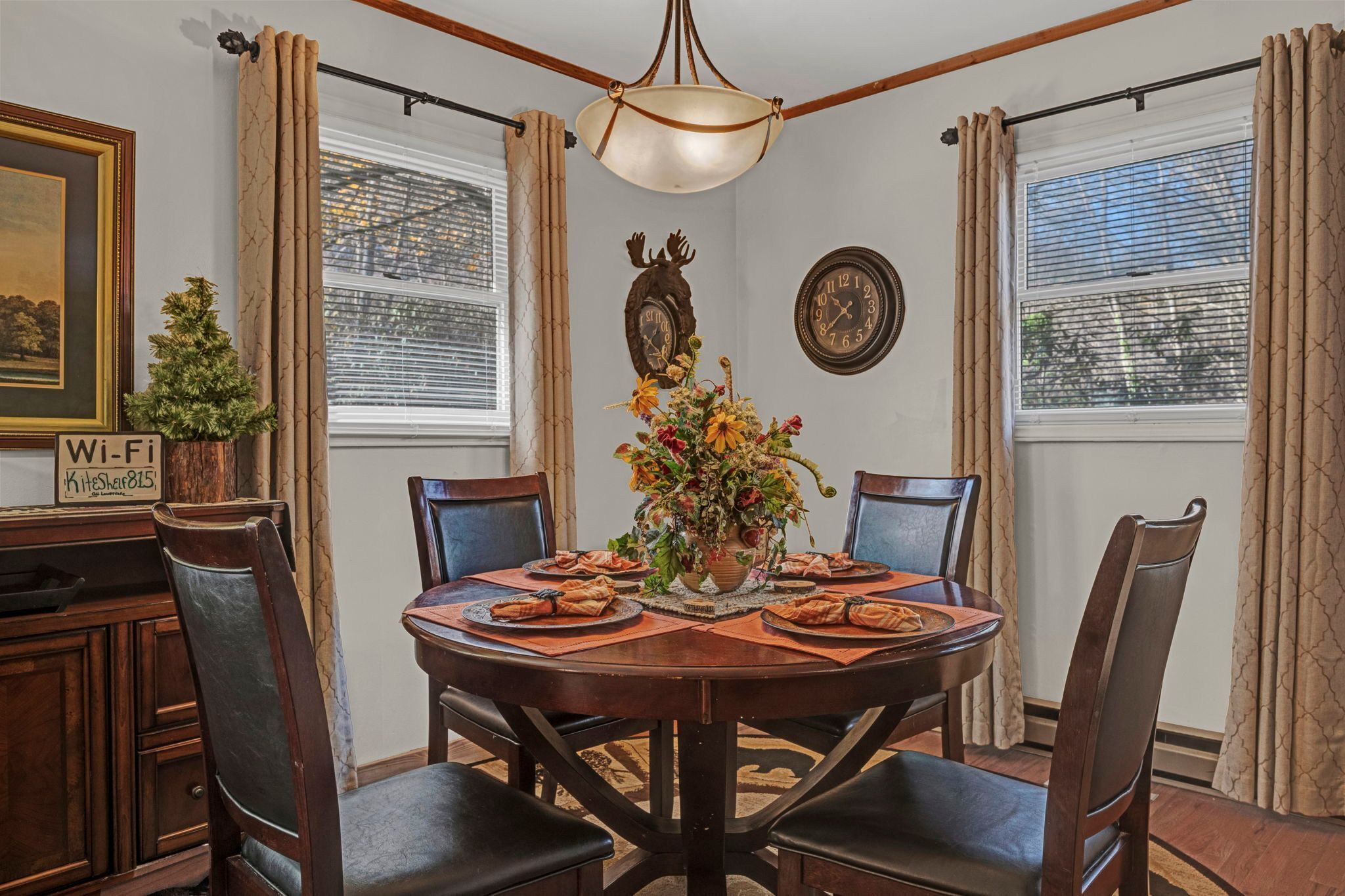 A dining room with a round table, chairs, a chandelier, a floral centerpiece, and a clock on the wall.