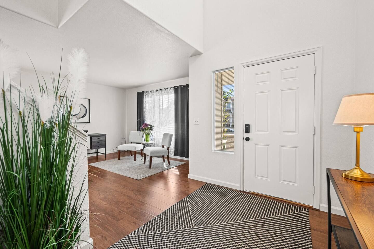 A modern living room with a wooden floor, a white door, a dining area with a table and chairs, and a lamp on a side table.