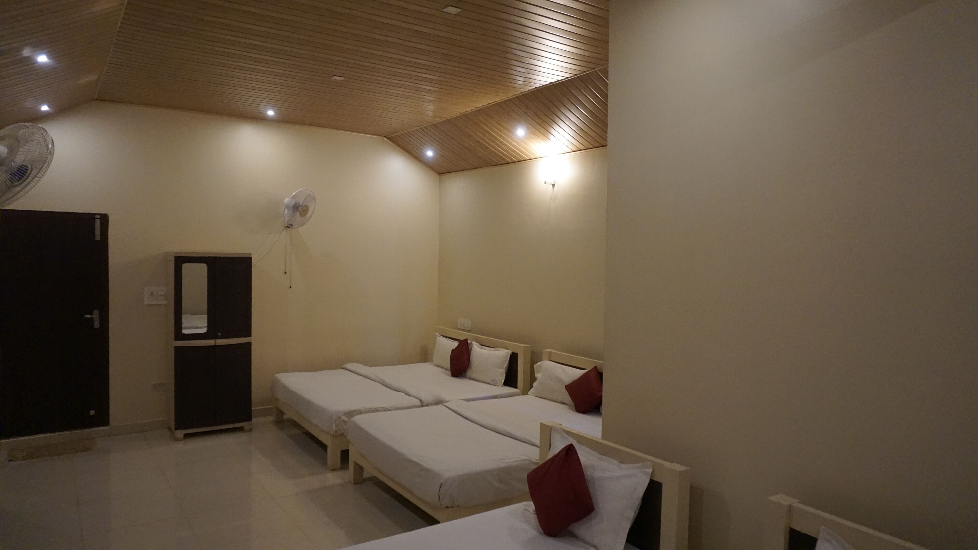 Una habitación de hotel con tres camas individuales, un armario de color oscuro, un ventilador de pie y un ventilador de techo.