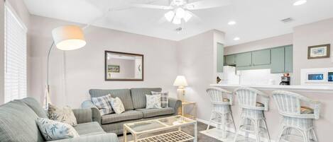 A living room with a sofa, coffee table, and wicker chairs. There is a ceiling fan, a lamp, and a kitchen area with a counter and cabinets.