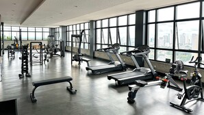 Un gimnasio moderno con cintas de correr, máquinas de pesas y un banco. Amplias ventanas ofrecen vistas a la ciudad.
