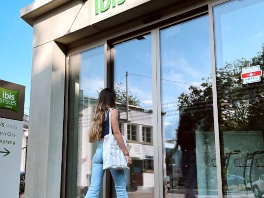 A hotel entrance with a glass facade, a sign indicating 'ibis Styles Hotel,' and a directional sign for 'Bern City Empfang.'