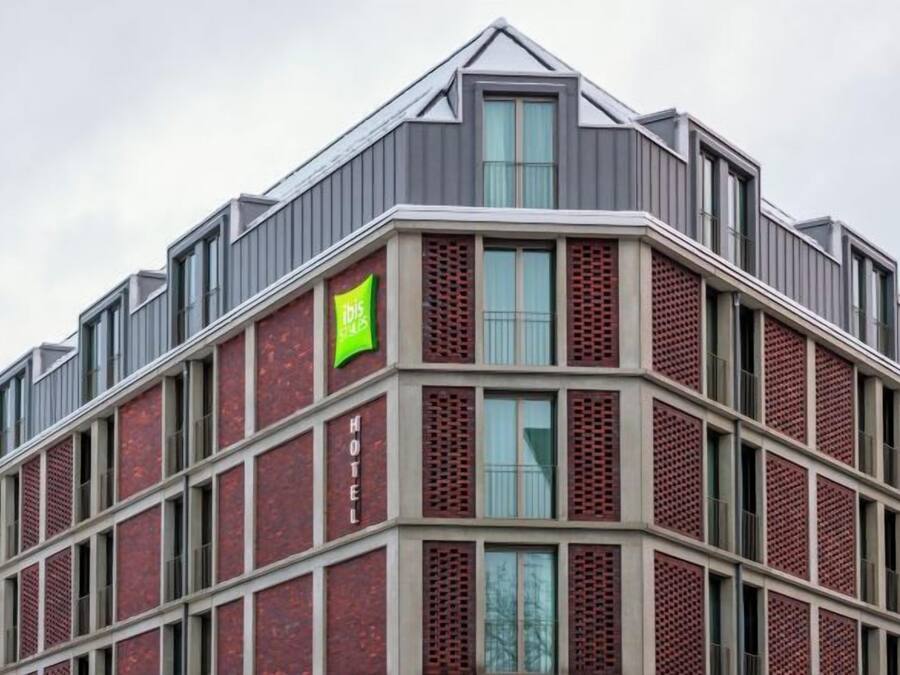 A modern hotel with a distinctive red brick facade, large windows, and a green sign indicating the name.