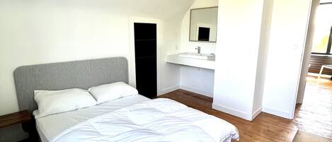 Een slaapkamer met een bed, nachtkastje, inbouwkleerkast, badkamer met wastafel en spiegel, en een houten vloer.