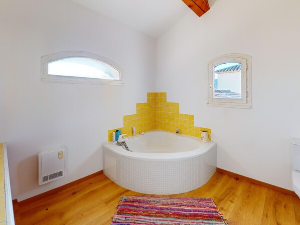A bathroom with a corner bathtub, yellow-tiled walls, a wooden floor, and a colorful rug.