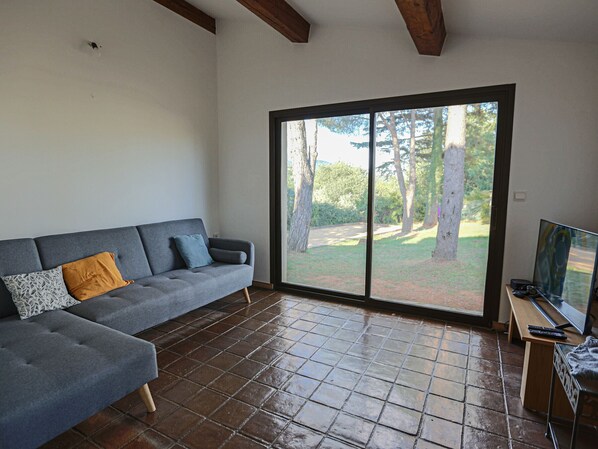 Un soggiorno con un divano grigio, una TV su un mobile in legno e un'ampia porta scorrevole in vetro che dà su un giardino.