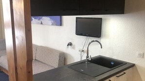 Cocina compacta con encimera negra, un lavamanos y armarios de madera. Un televisor de pantalla plana está montado sobre el lavamanos.