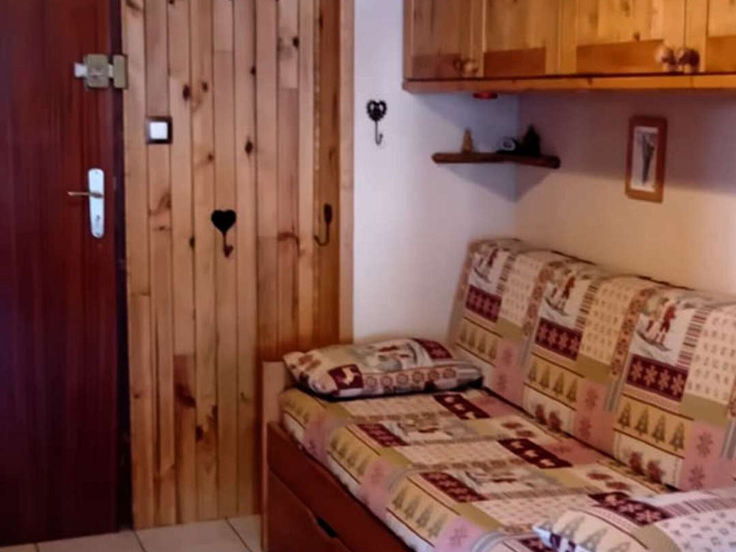 Een knus, klein kamertje met een houten garderobe, een bed met beddengoed met een patroon en een klein plankje met decoratieve spullen.
