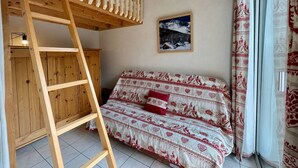 A bunk bed with a wooden ladder, a sofa with patterned bedding, a framed picture on the wall, and a window with curtains. - Studio Cabin 5 pers, Parking, Near Slopes (Briançon)