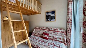 A bunk bed with a wooden ladder, a sofa with patterned bedding, a framed picture on the wall, and a window with curtains.