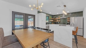 A modern kitchen with a central island, a dining area with a wooden table, and a view of the outdoors through large windows. - Hot Tub | Speakeasy | Basketball Court | Sleeps 12 (Cedar Park)