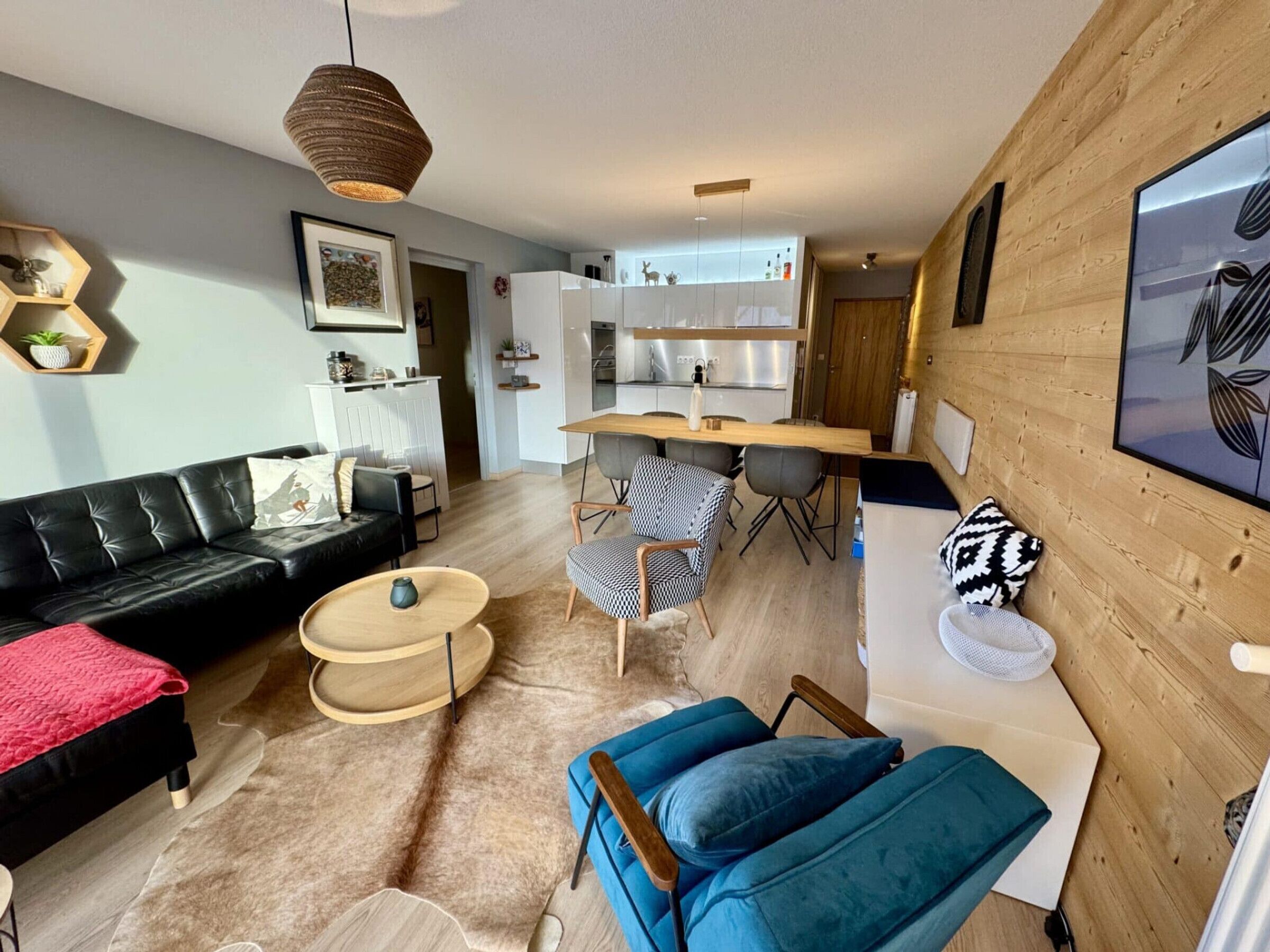 A modern living room with a black leather sofa, a blue armchair, and a wooden coffee table. A kitchen with white cabinets and a dining area with a wooden table and chairs is visible in the background.
