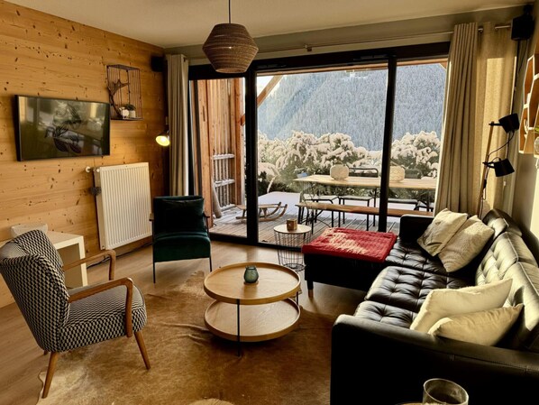 A cozy living room with a leather sofa, a round coffee table, and a chair. A large window offers a view of a snowy mountain.