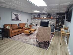 Un salon avec un canapé marron, un fauteuil à motifs, une table basse, une cheminée et une bibliothèque.