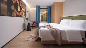 A modern hotel room with a large bed, a desk with a chair, a small table with a vase, and a large framed picture on the wall.