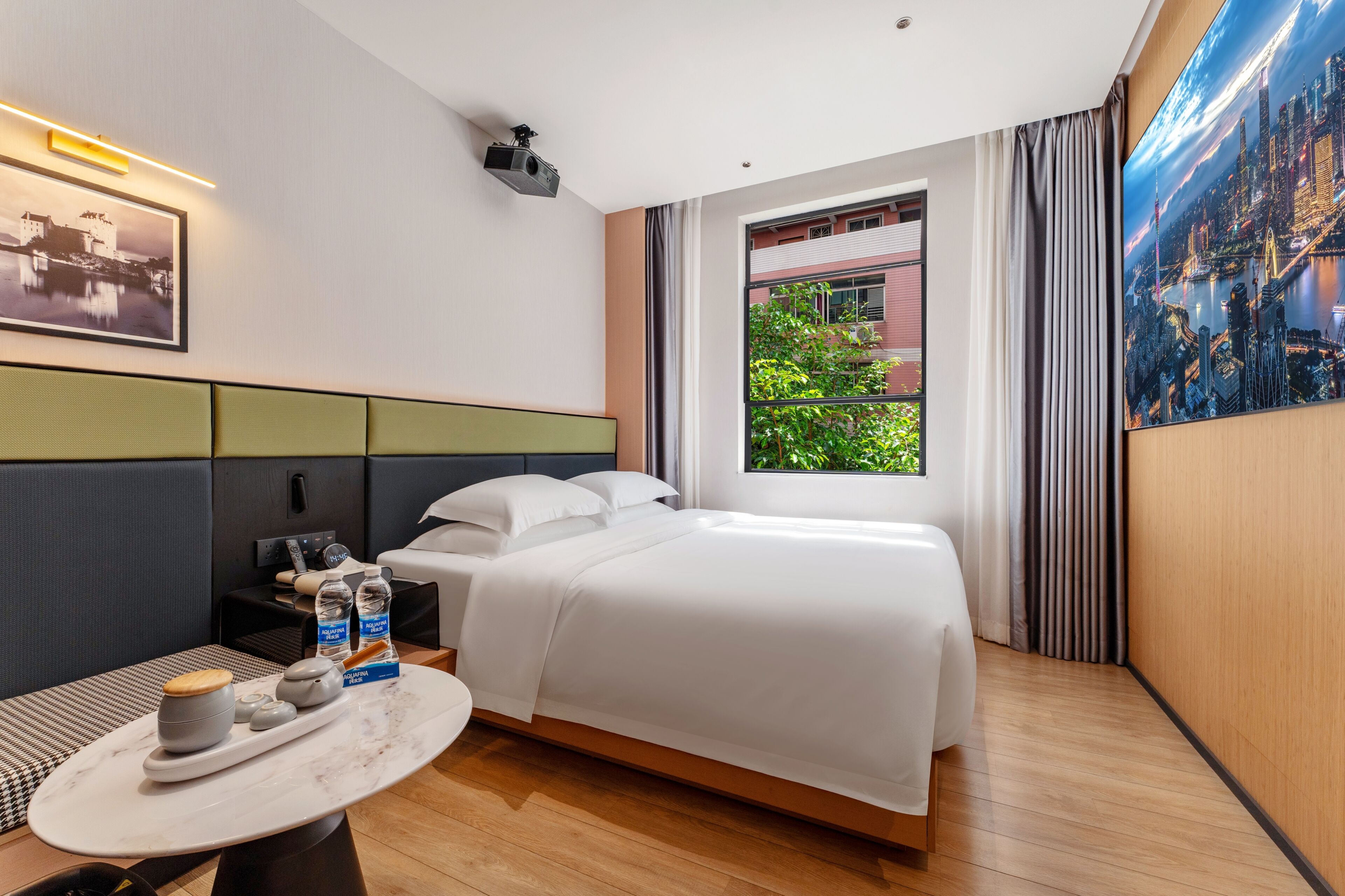 A hotel room with a bed, a desk with a chair, a small table with a cup and bottles, a projector, and a large window with a cityscape view.