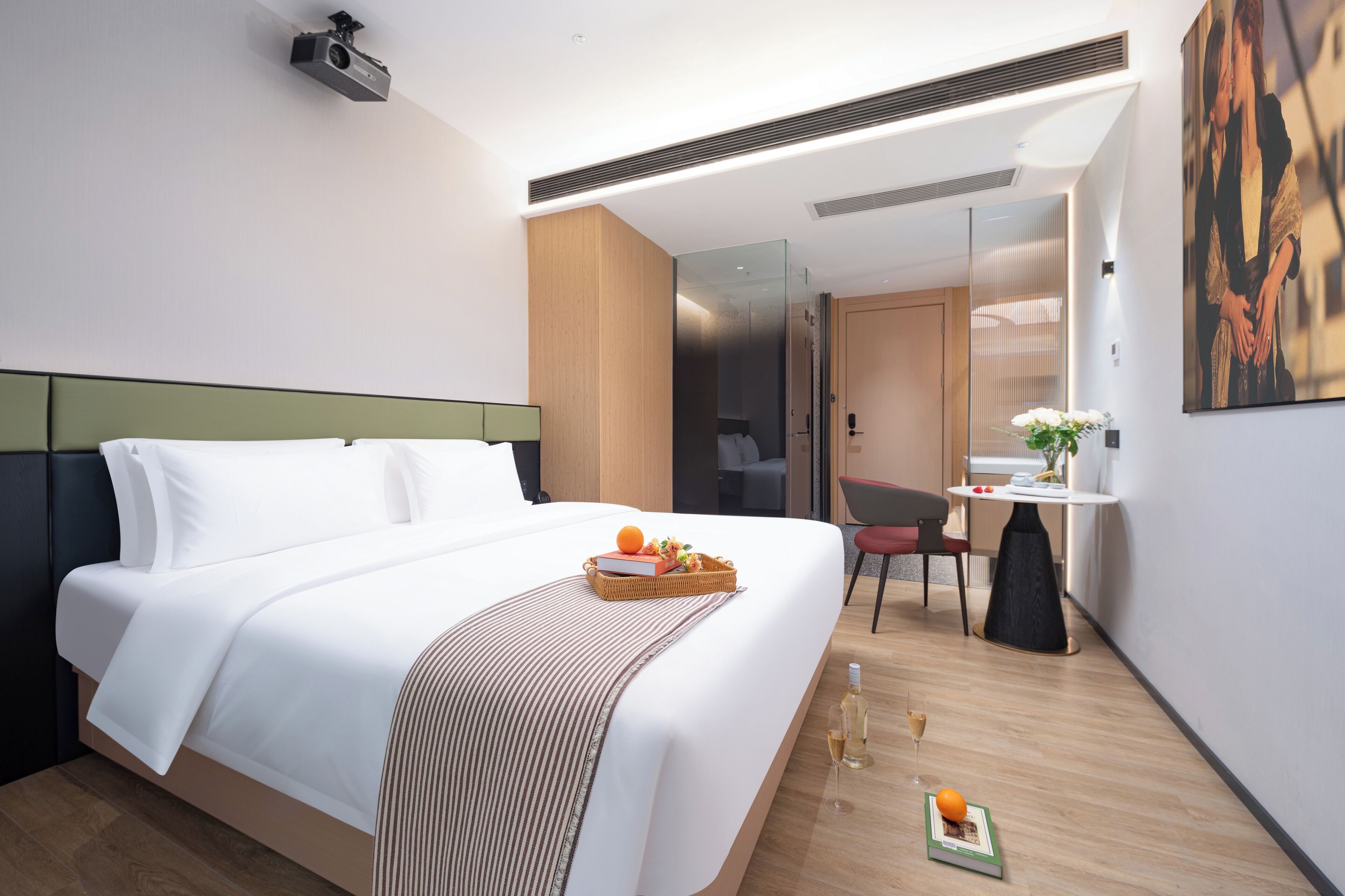 Ein modernes Hotelzimmer mit einem aufgeräumten Bett, einem kleinen Schreibtisch mit Stuhl, einer Glastrennwand und einem Bild an der Wand.