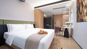 Ein modernes Hotelzimmer mit einem aufgeräumten Bett, einem kleinen Schreibtisch mit Stuhl, einer Glastrennwand und einem Bild an der Wand.