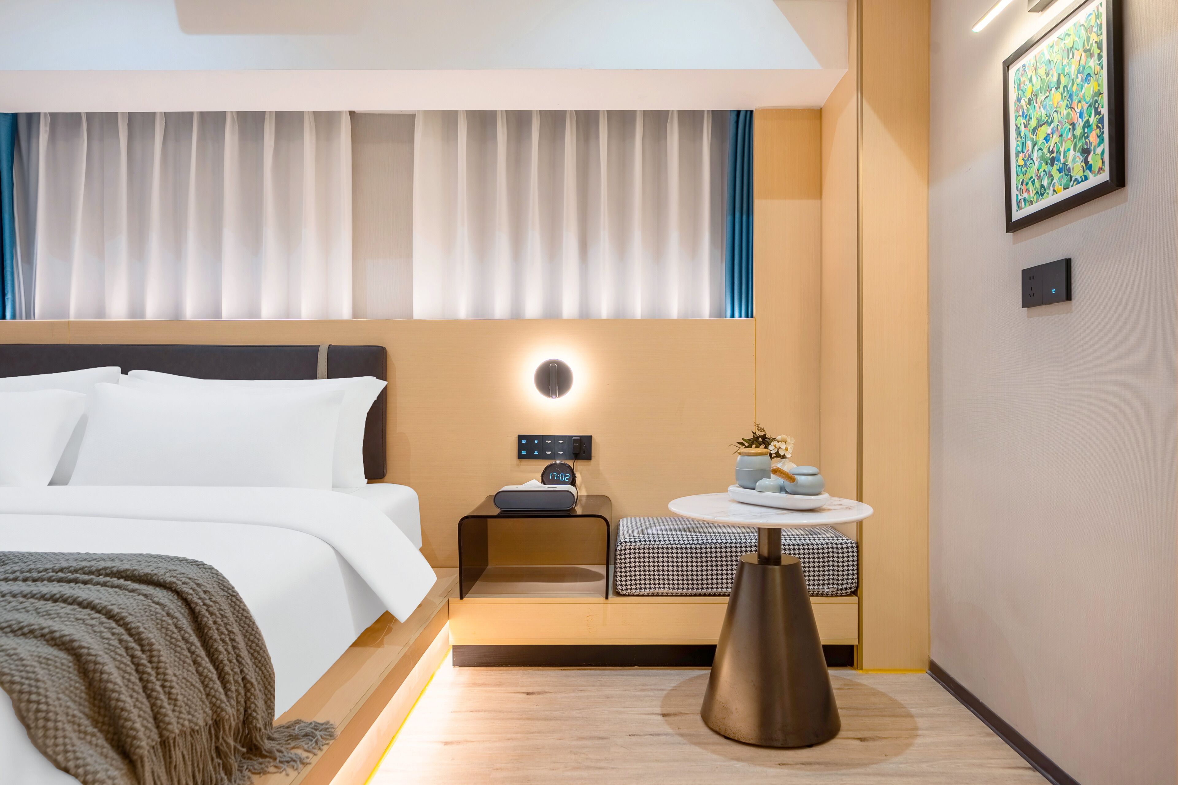 Una habitación de hotel moderna con una cama bien tendida, una mesita de noche con un reloj, una pequeña mesa redonda con objetos decorativos y una lámpara de pared.