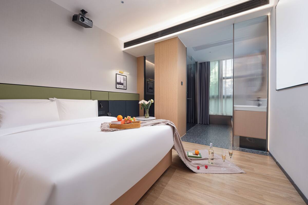 Une chambre d’hôtel avec un grand lit, une petite table sur laquelle se trouvent des fruits et une bouteille, et une vue sur le couloir.