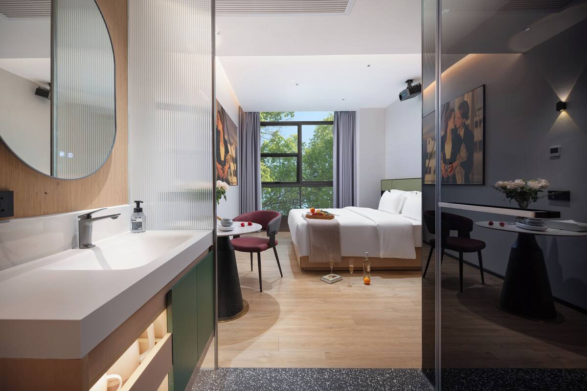 Une chambre d’hôtel moderne avec un grand miroir, un lavabo rond, un lit avec une tête de lit blanche, une petite table avec un vase et une fenêtre avec des rideaux.