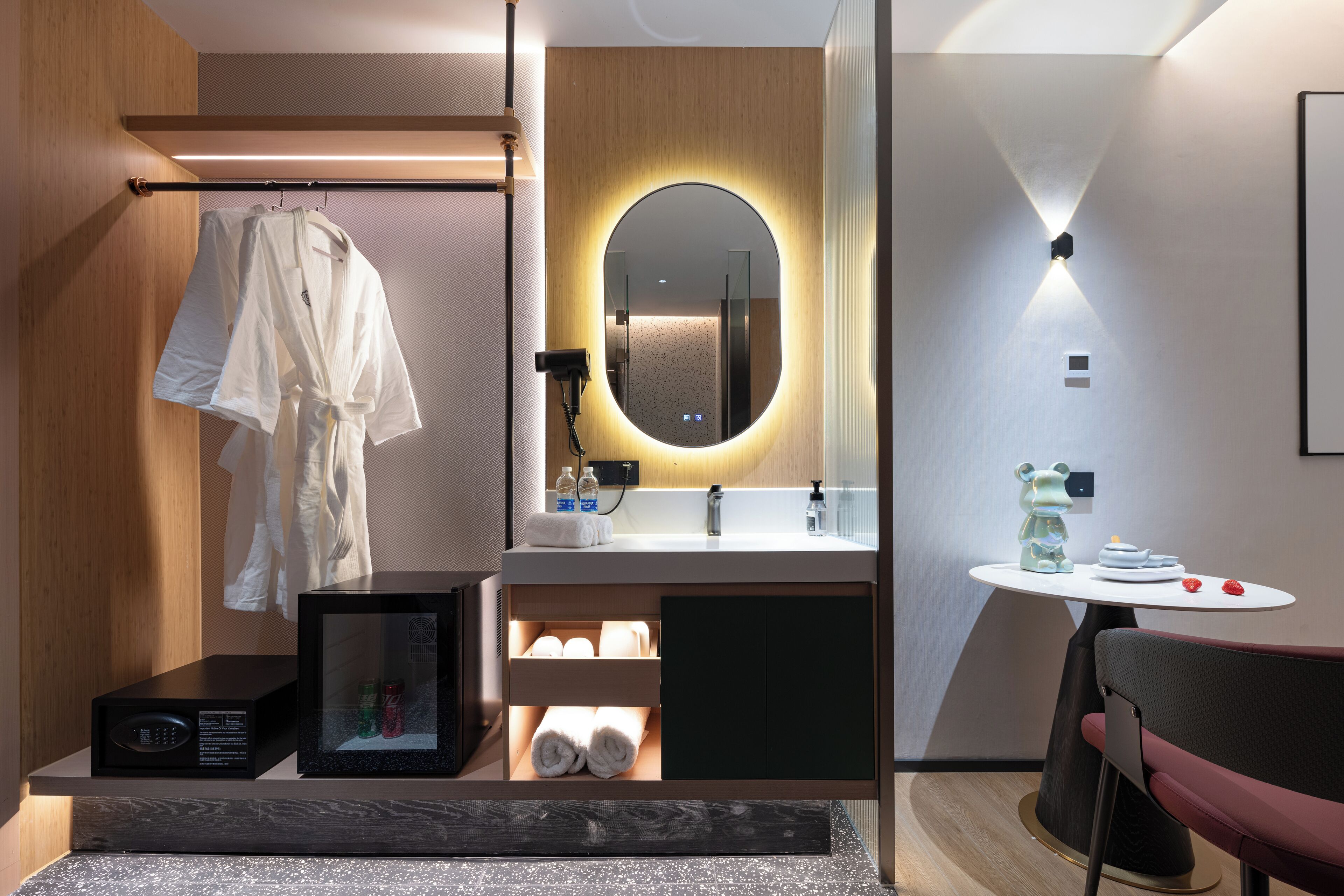 Un baño moderno con tocador de madera, un espejo redondo con iluminación inferior, una mesita con artículos de aseo y una silla.