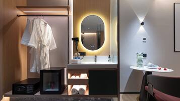 Un baño moderno con tocador de madera, un espejo redondo con iluminación inferior, una mesita con artículos de aseo y una silla.