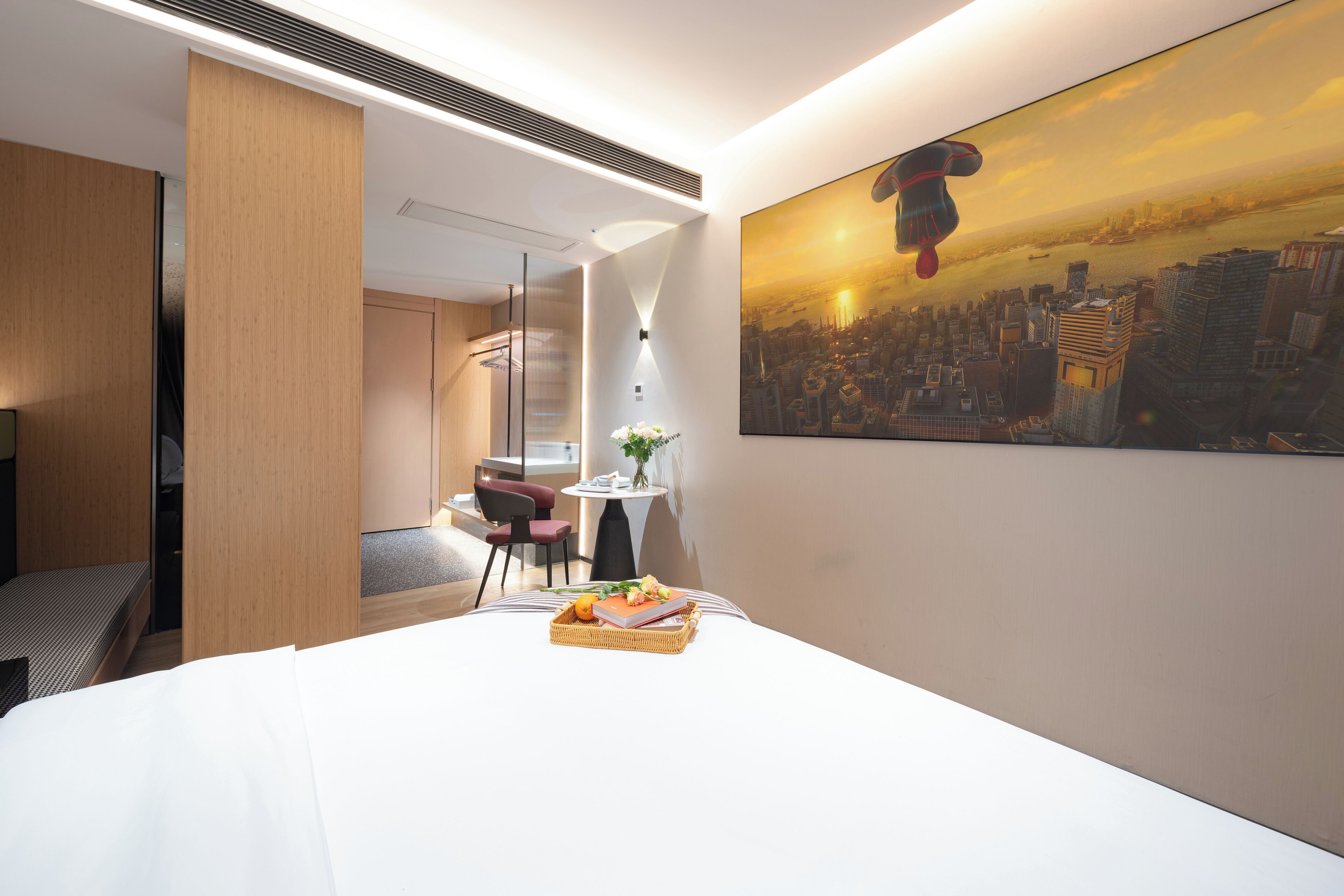 Quarto de hotel com uma cama, uma mesa de jantar com uma cesta de frutas, uma cadeira, uma pequena mesa com um vaso e uma pintura de paisagem urbana na parede.