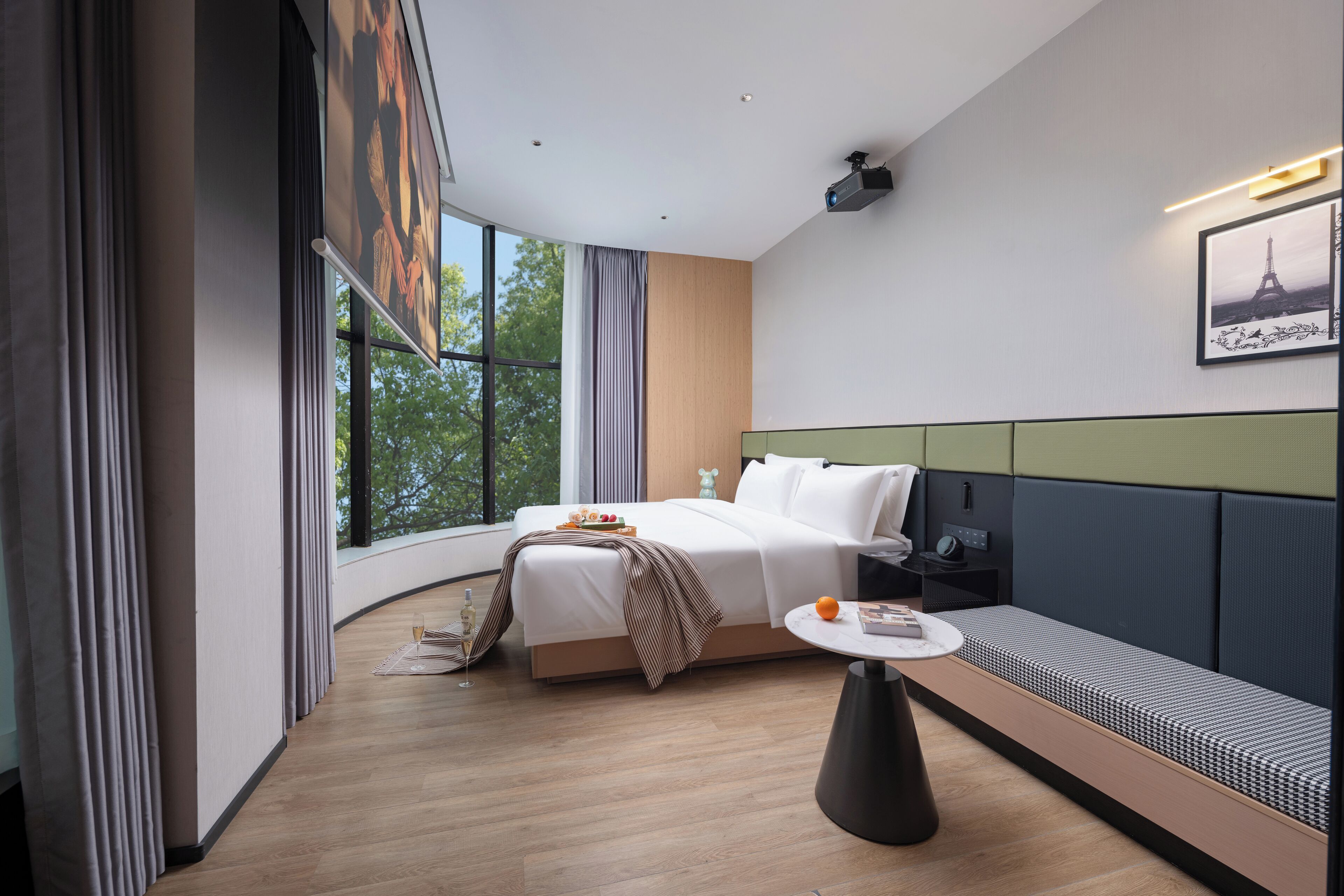 A modern hotel room with a large bed, a seating area, a small table, and a large window offering a view of trees.