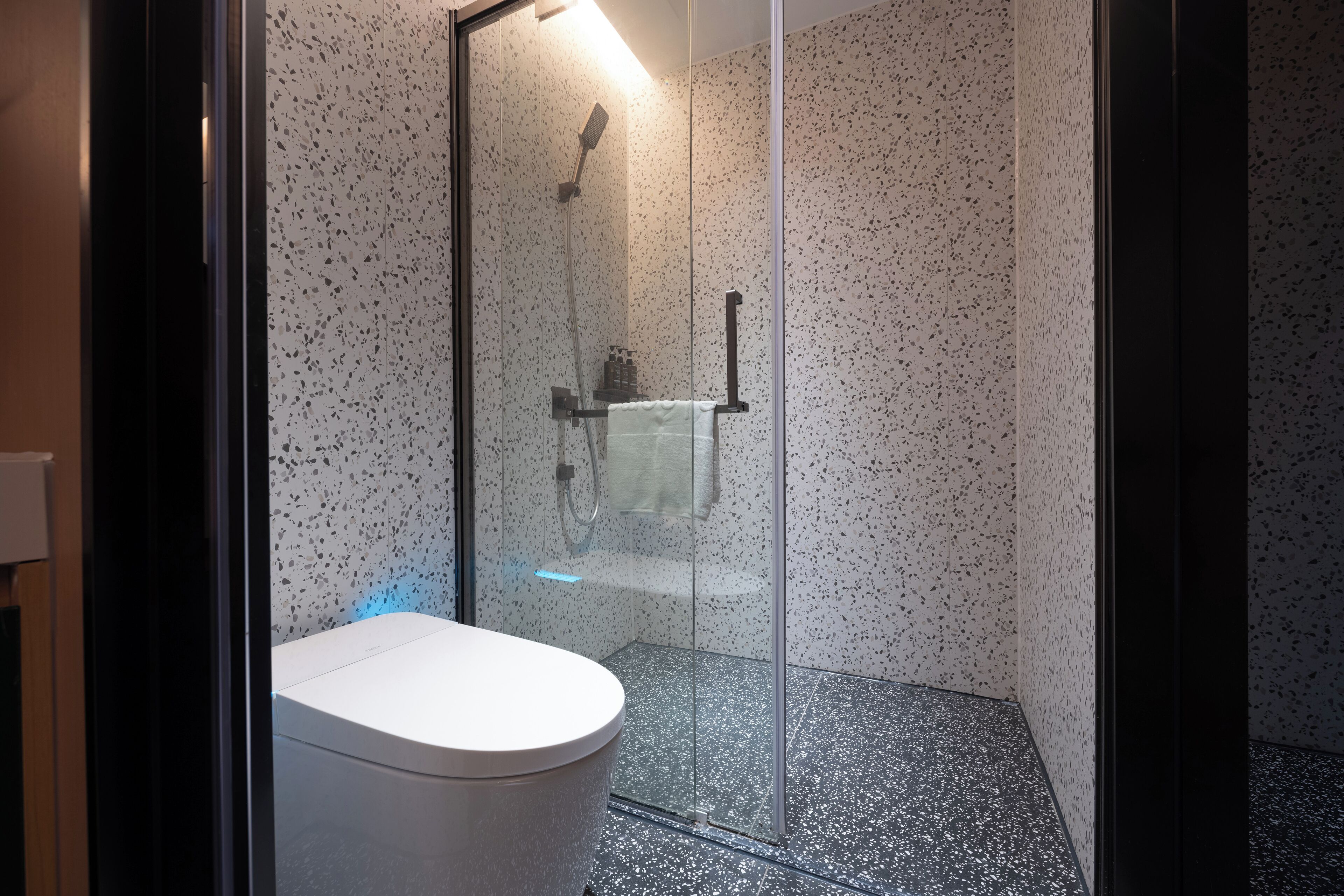 Un baño moderno con ducha envuelta en vidrio, inodoro blanco y baldosas de suelo con motas.