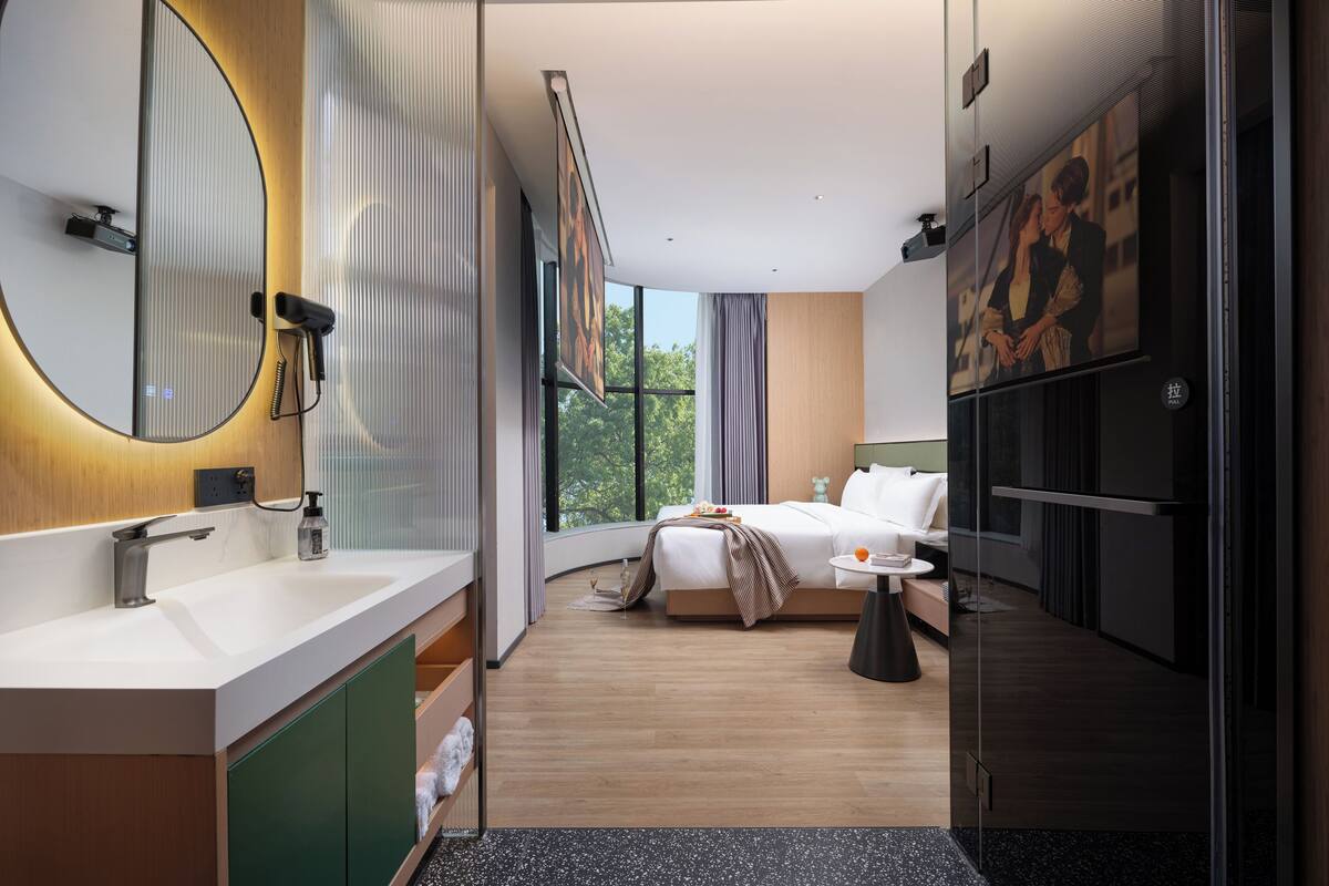 Une chambre d’hôtel moderne dotée d’un grand lit, d’une salle de bain séparée avec un meuble-lavabo et d’une grande fenêtre laissant entrer la lumière naturelle.
