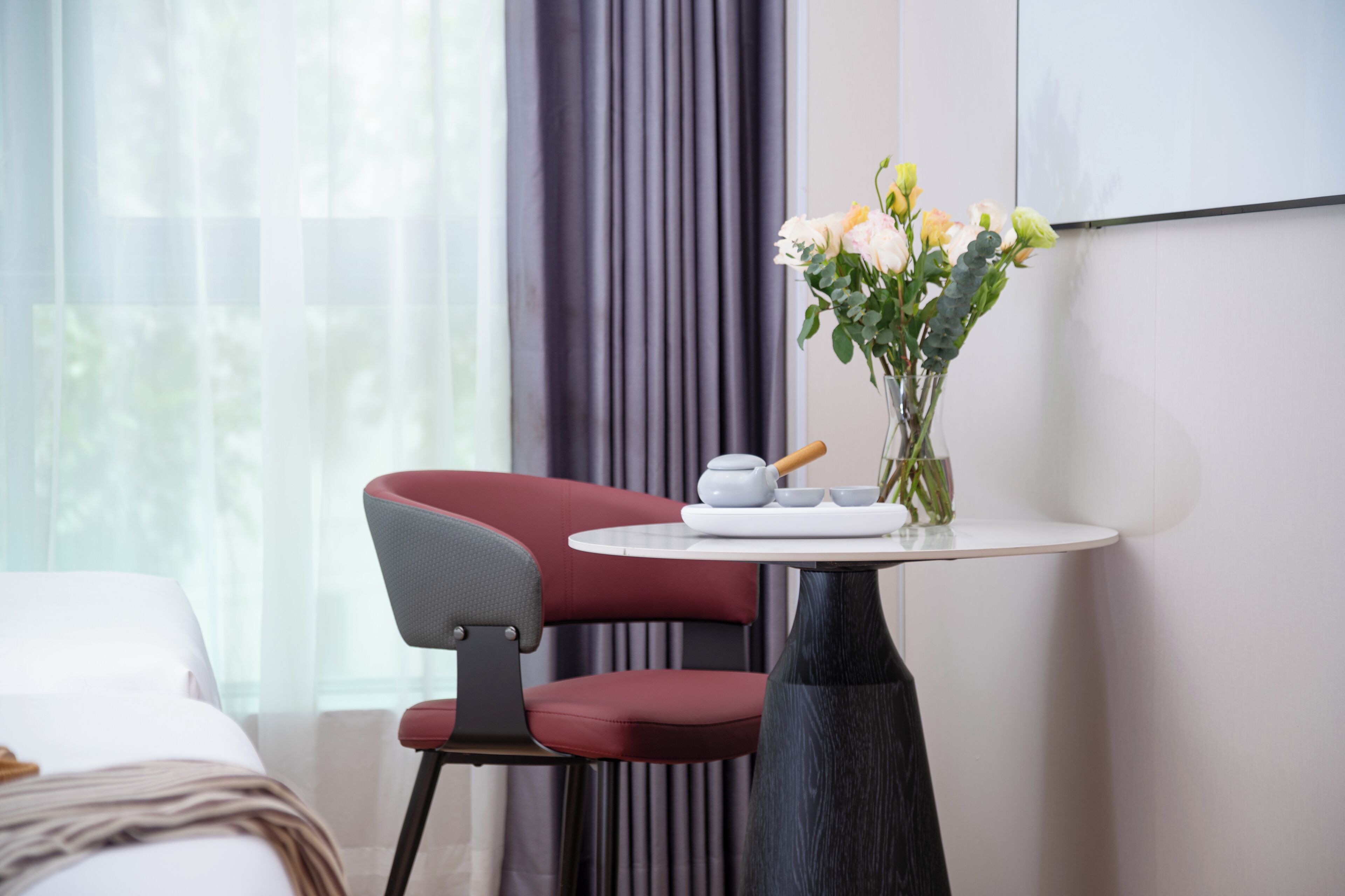 A hotel room with a bed, a red chair, a small round table with a vase of flowers, and a wall-mounted screen.