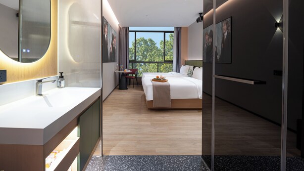 A modern hotel room with a large bed, a separate bathroom with a large mirror, and a small seating area with a table and chair.